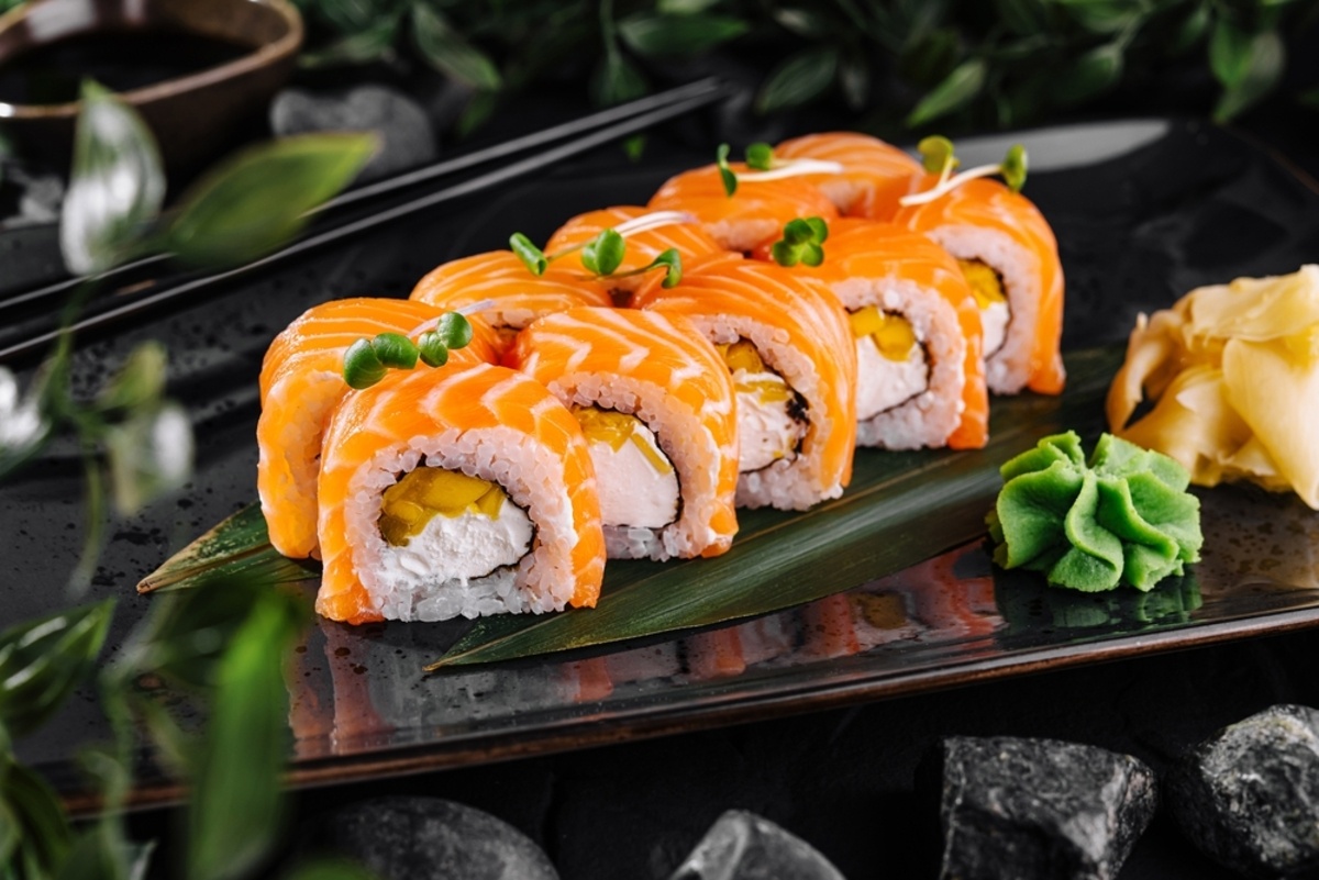 Freshly prepared sushi rolls topped with salmon, garnished with microgreens and served with wasabi and pickled ginger on a dark slate board
