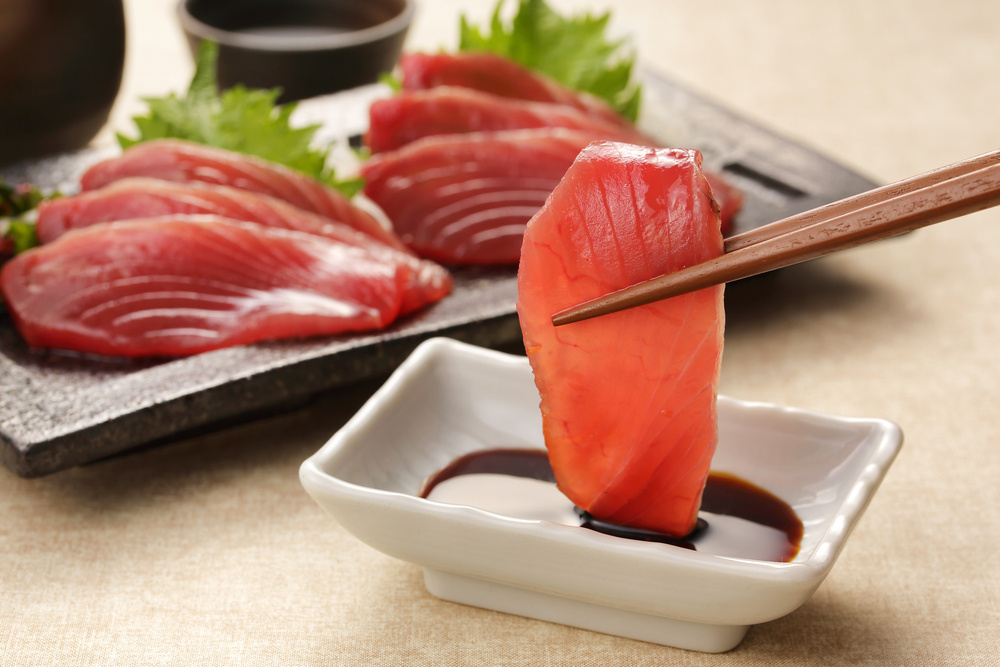 Fresh tuna sashimi in Brooklyn NY held by chopsticks over soy sauce with a platter of sliced fish in the background