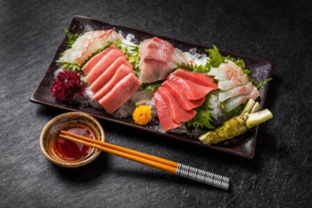 A traditional sashimi platter featuring tuna yellowtail and white fish cuts over ice and shiso leaves served with soy sauce chopsticks and fresh wasabi root