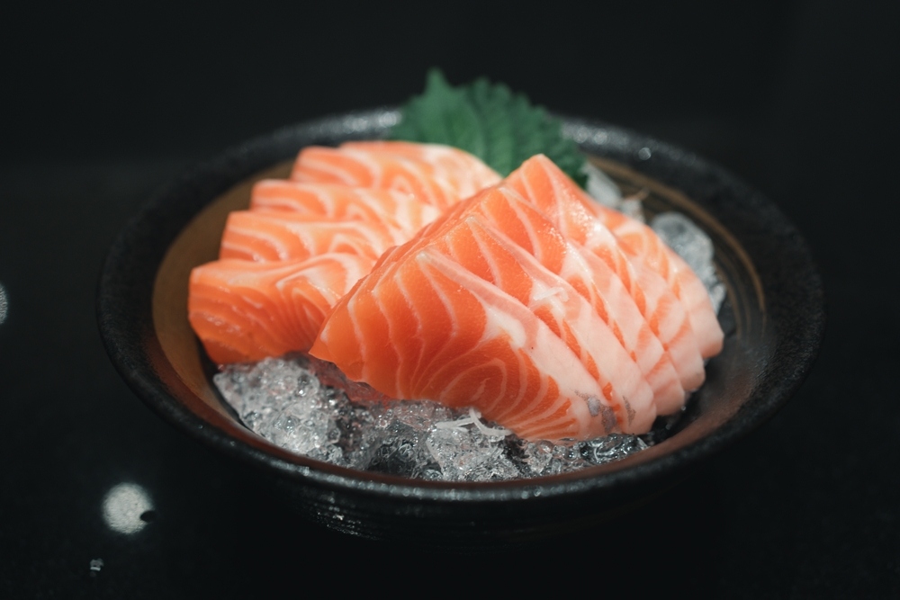 Thick-cut salmon sashimi grade slices served over ice in a dark ceramic bowl with fresh shiso garnish