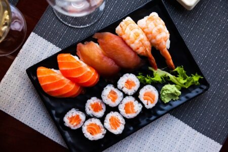 An overhead view of a mixed sushi texture plate featuring salmon sashimi slices tuna and shrimp nigiri and salmon hosomaki rolls served with wasabi on a black dish