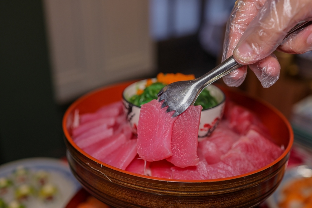 Chef using tongs to plate fresh tuna sashimi in Coney Island Brooklyn served in a traditional Japanese wooden bowl with seaweed and garnishes at Masuta