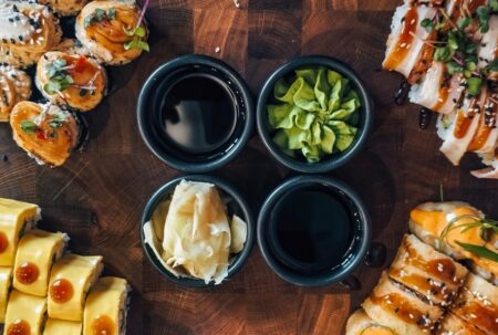 Overhead view of a sushi spread showing how to order sushi with soy sauce wasabi and pickled ginger condiment bowls surrounded by assorted specialty rolls on a wooden board