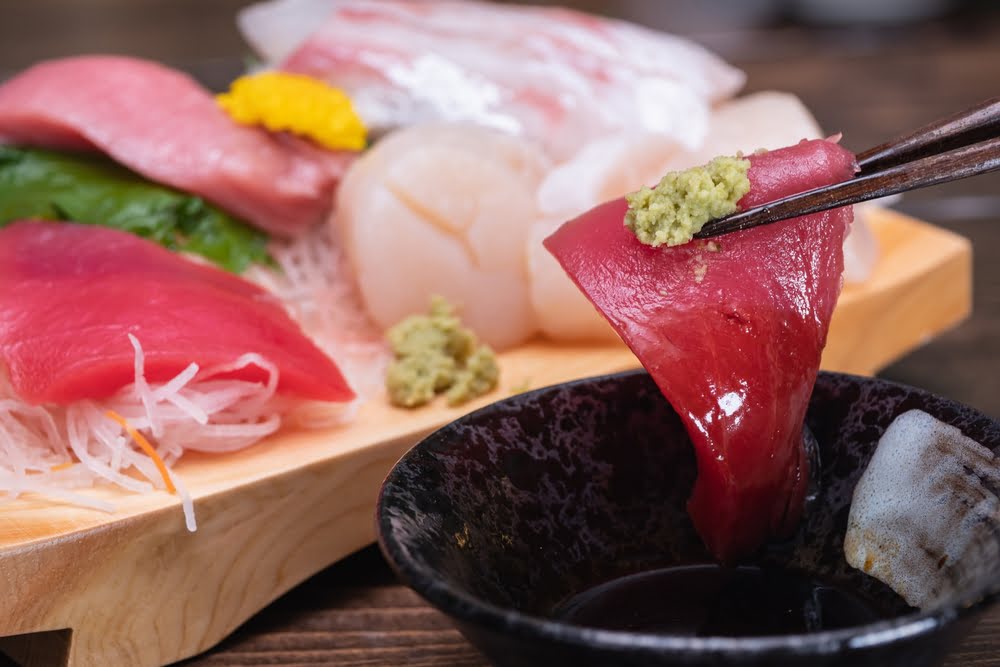 A close-up showing how to taste sashimi properly as chopsticks lift a fresh tuna slice topped with wasabi over a soy sauce bowl with a multi-species sashimi board in the background