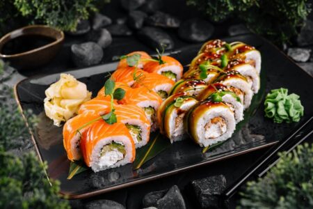 Two Japanese sushi culture inspired specialty roll varieties on a dark ceramic plate featuring salmon-topped uramaki with microgreens alongside avocado rolls drizzled with teriyaki sauce and sesame seeds