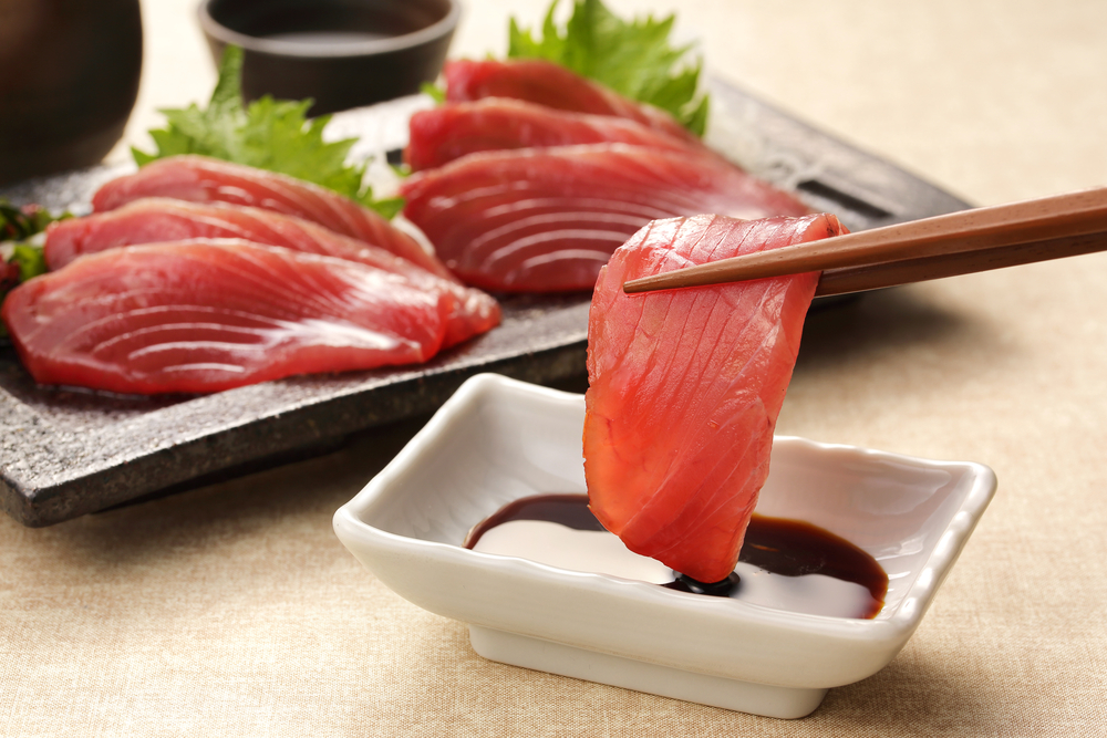 Fresh tuna sashimi in Gravesend Brooklyn held by chopsticks above a soy sauce dish with a full platter of hand-sliced tuna and shiso leaves served at Masuta