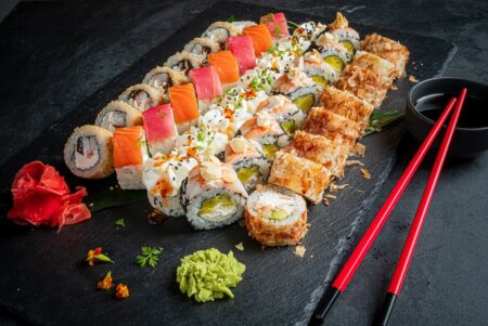 A large spread of specialty sushi rolls on a dark slate board featuring rainbow rolls crispy tempura rolls cream cheese rolls and sauce-drizzled uramaki served with wasabi and pickled ginger