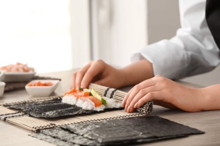 A sushi chef perspective captured as a chef rolls a maki sushi roll on a bamboo mat with salmon cucumber and sushi rice on nori sheets at a professional kitchen counter