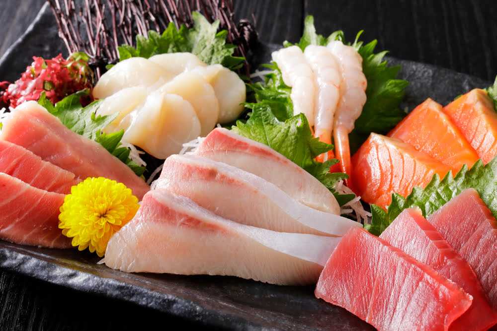 A great sashimi restaurant platter featuring scallop shrimp yellowtail tuna and salmon arranged over shiso leaves and shredded daikon with an edible flower garnish on a dark slate board